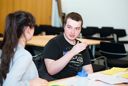 Young man gestures as he speaks to peer in a classroom